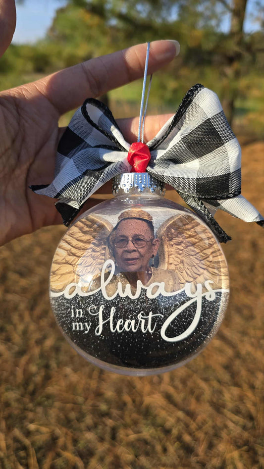 Memorial ornament with photo of a person wearing angel wings and text 'Always in my Heart' held by a hand outdoors.